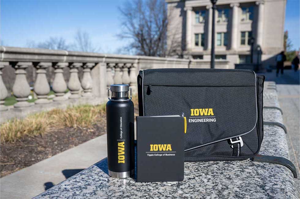Unit lockups displayed on a laptop bag, journal, and water bottle set against a backdrop of the Pentacrest.